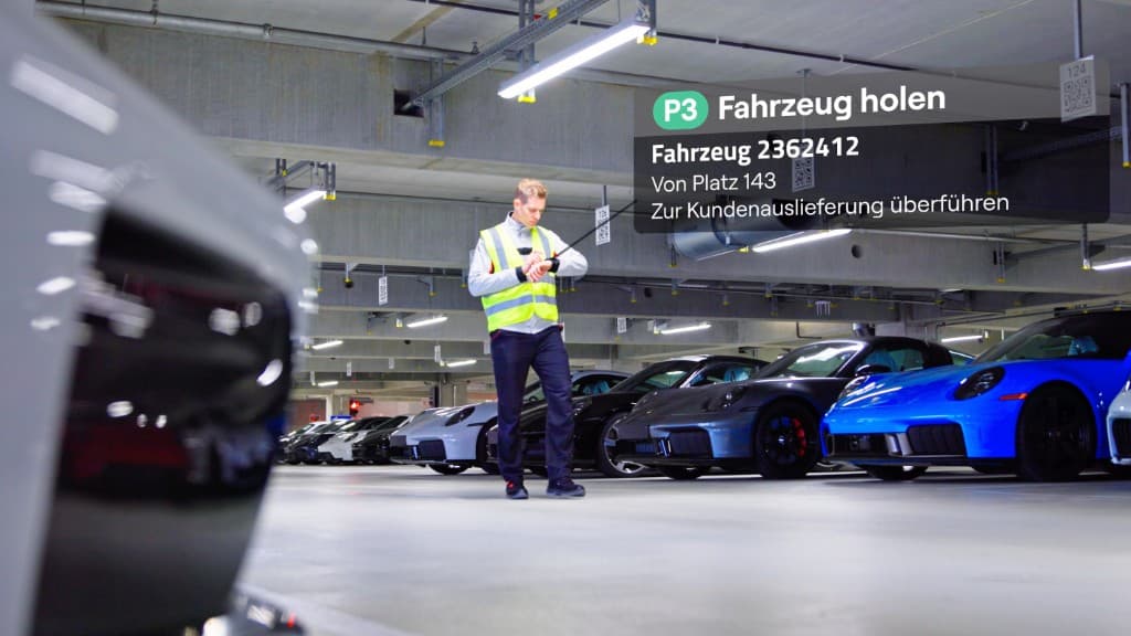 Technician in a high-visibility vest follows guided task instructions on an industrial smartwatch, with contextual task details shown in an augmented overlay beside vehicles in a connected facility